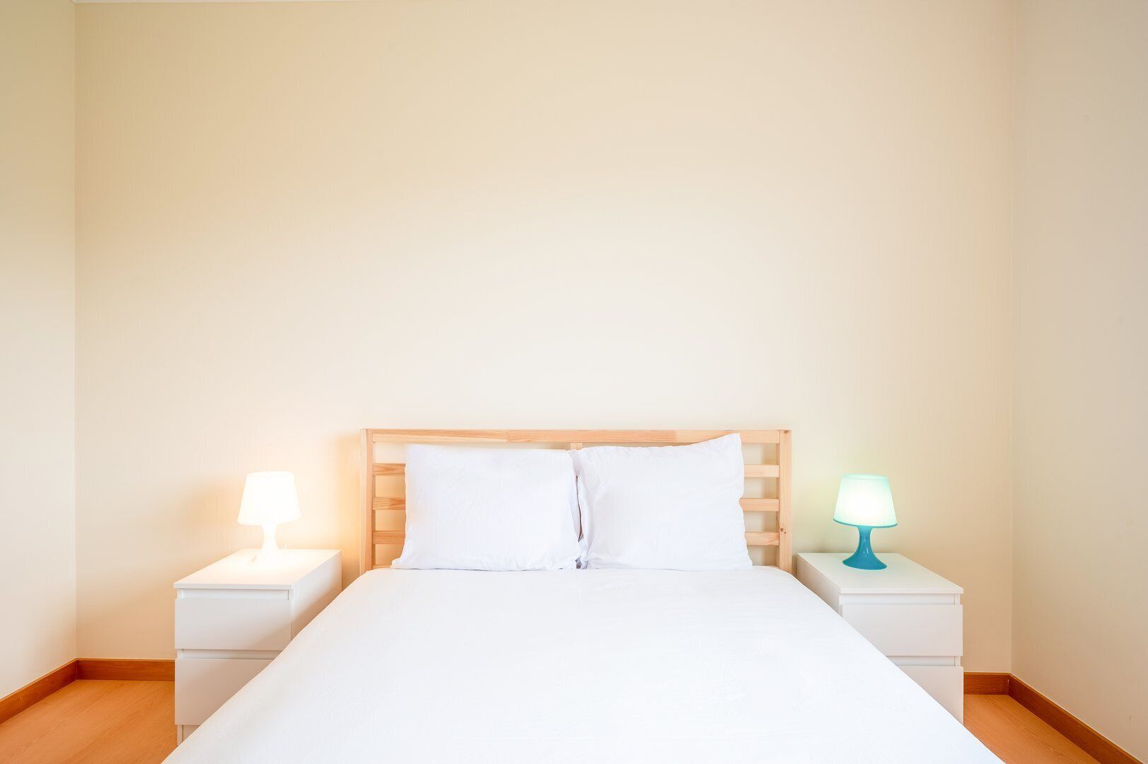 A neatly made bed with white bedding and a wooden headboard. There are two bedside tables, each with a lamp. The room has plain walls and a wooden floor.