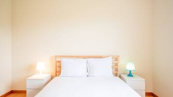 A neatly made bed with white bedding and a wooden headboard. There are two bedside tables, each with a lamp. The room has plain walls and a wooden floor.