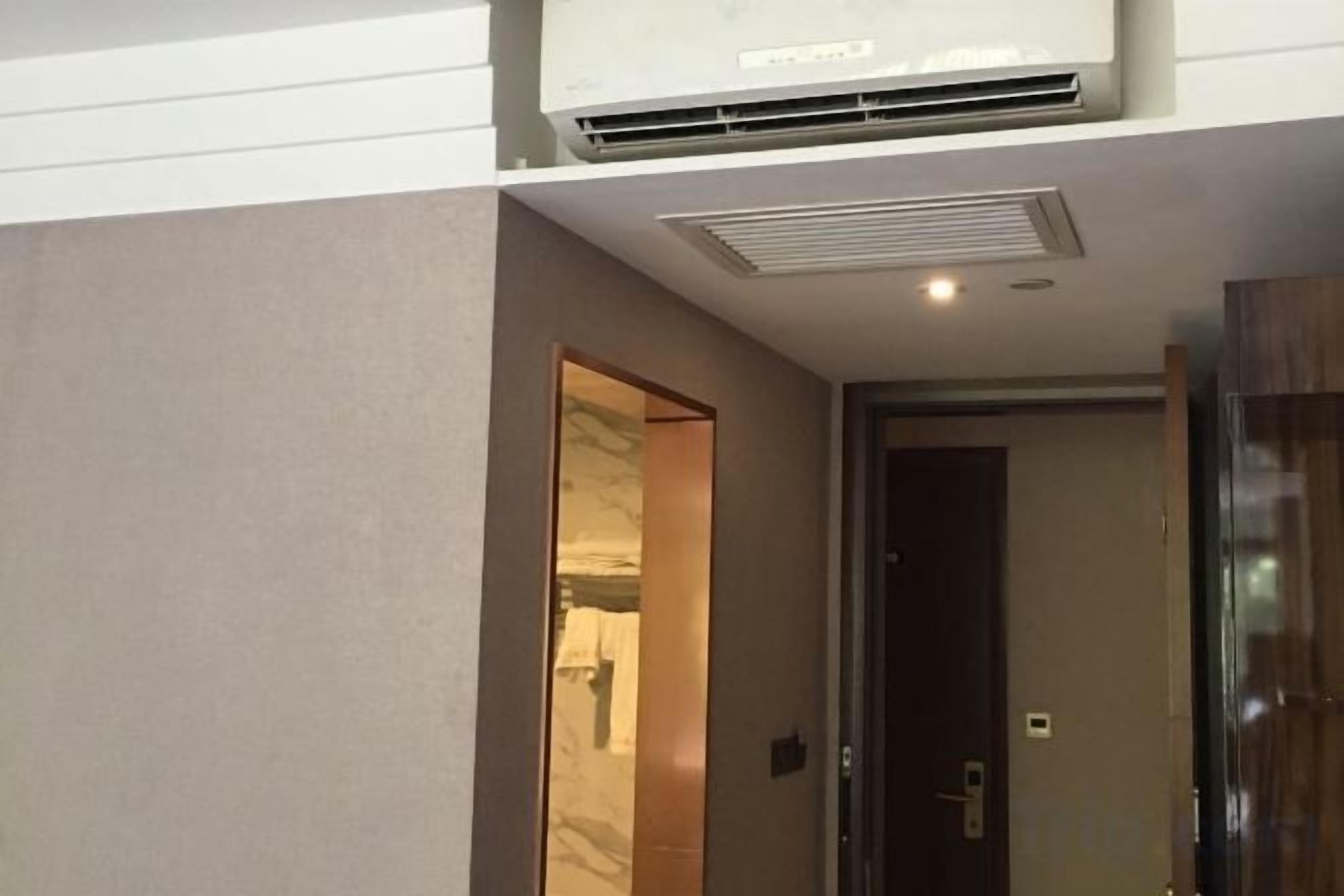 A hotel room with a ceiling-mounted air conditioner, a door leading to another room, and a visible light switch.