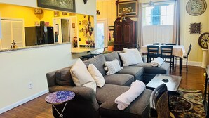 Un salon comprenant un canapé d’angle, une table à manger avec des chaises, un coin cuisine et une grande horloge murale.