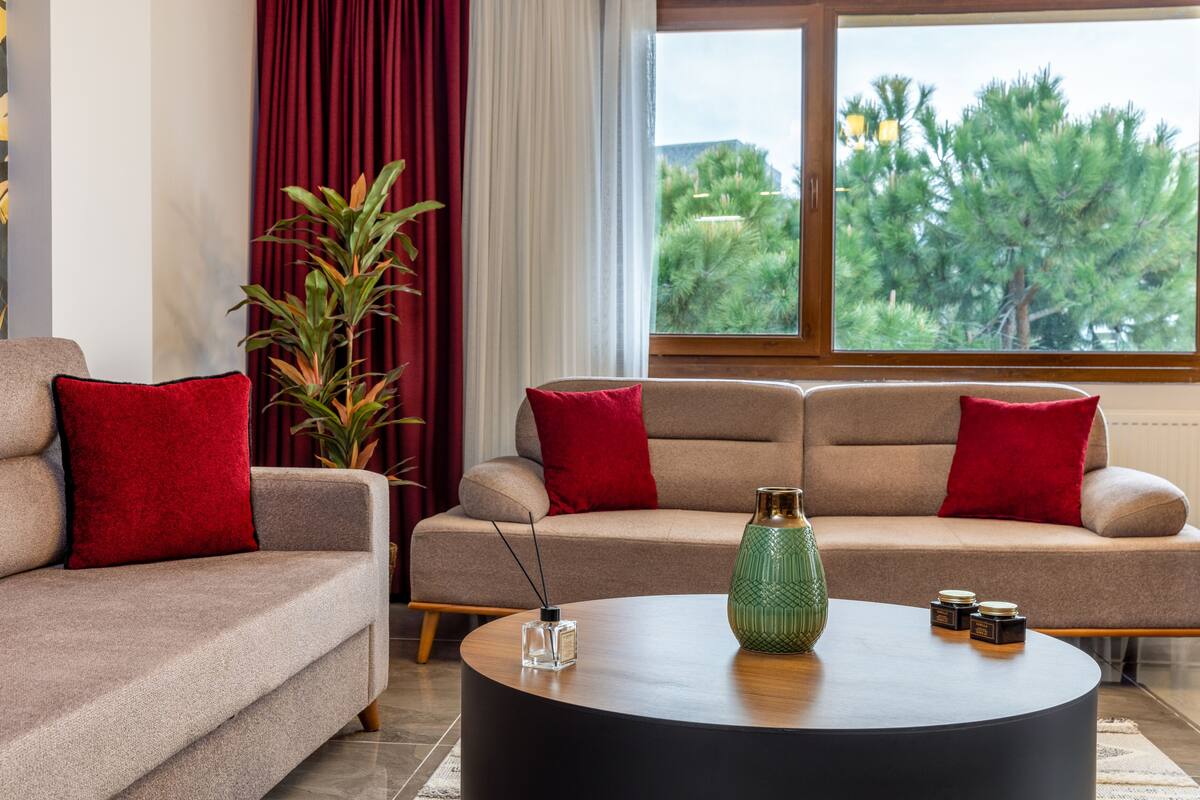 A modern living room with two sofas, red cushions, a round coffee table with decorative items, a large window with a view of trees, and a potted plant.