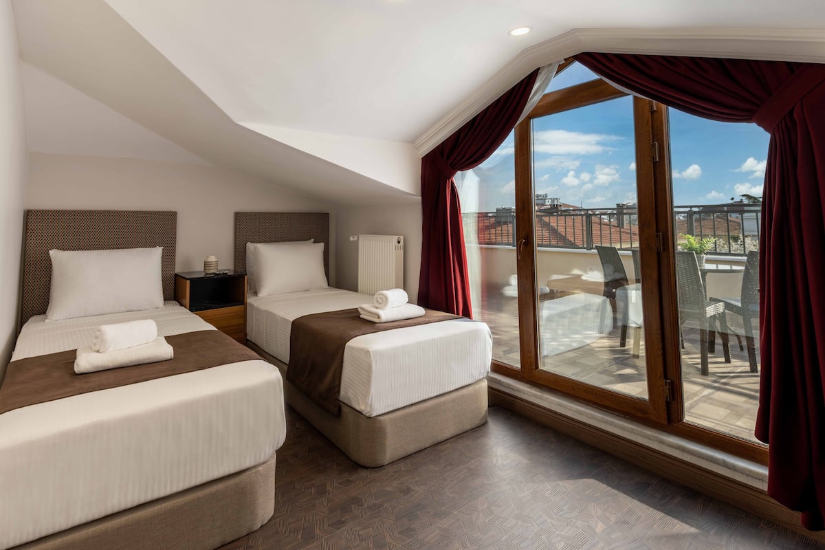 A hotel room with two beds, a large window with curtains, a view of the city, and a balcony with outdoor furniture.
