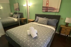 Two beds with grey bedding and geometric patterns, a framed picture of the Golden Gate Bridge, and a lamp and bottle on bedside tables.