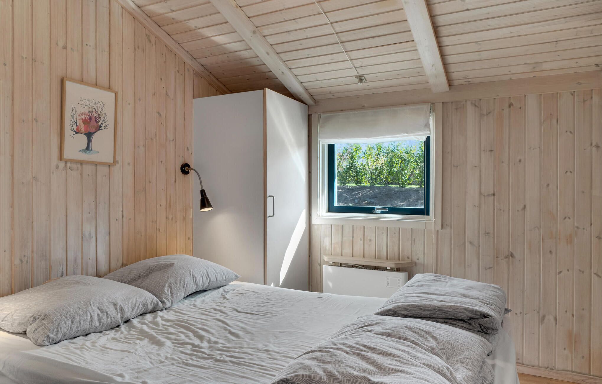 A bedroom with a wooden ceiling, a bed with white bedding, a nightstand, a desk, a window with a view of trees, and a wall-mounted lamp.