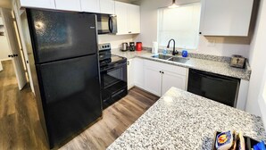 A compact kitchen with a black refrigerator, microwave, stove, and dishwasher. It features white cabinets, a granite countertop, and a window with blinds. - Astor Serene Riverfront Retreat with 2 boat slips and a dock (Astor)
