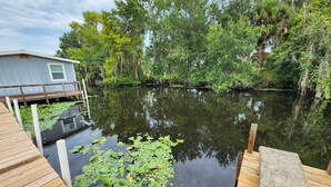 A wooden deck with a dock extending into a calm body of water surrounded by lush greenery and trees. - Astor Serene Riverfront Retreat with 2 boat slips and a dock (Astor)