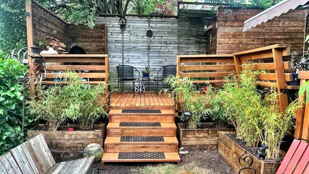 Una terraza de madera con un banco, una mesa pequeña con sillas y una escalera de madera que conduce a un área techada.