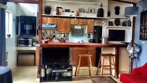 Cocina compacta con armarios de madera, microondas y televisor. Hay taburetes y una silla roja en la sala.