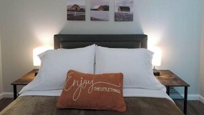 En pænt redt seng med en pude, der står "Nyd de små ting". Tre indrammede billeder hænger på væggen over sengen.