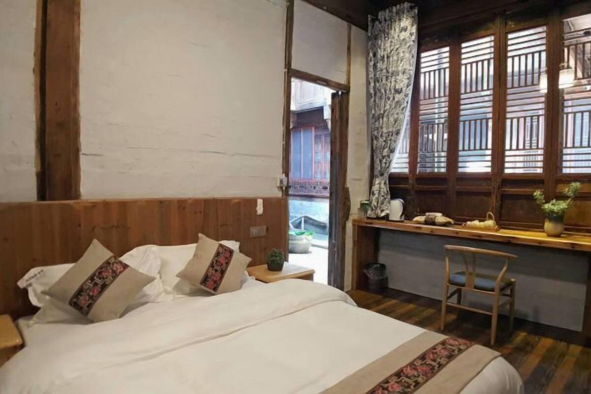 A bedroom with a wooden headboard, a bed with white bedding and patterned pillows, a nightstand, a chair, a desk with a plant, and a window with wooden shutters.