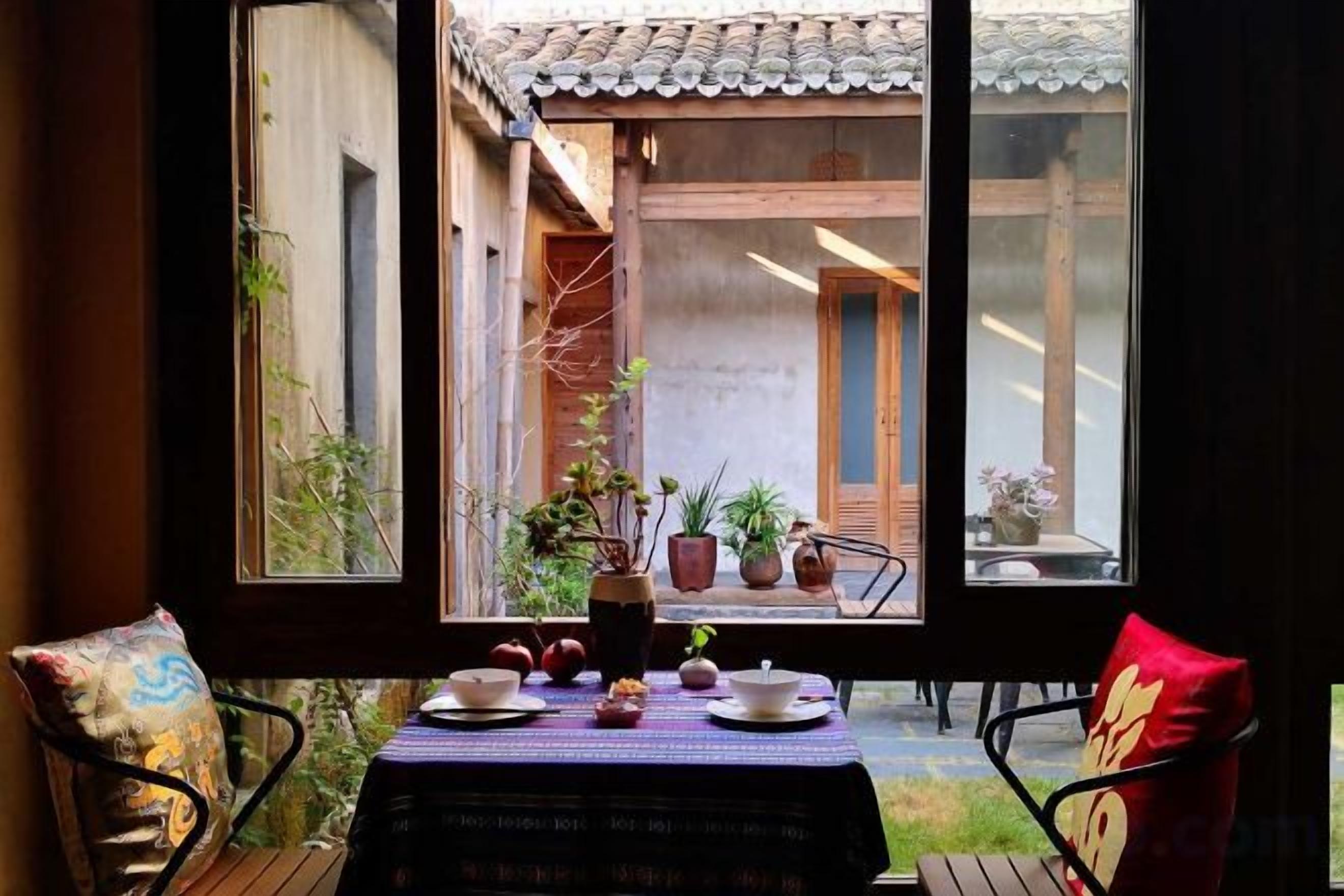 A dining area with a table set for two, a view of a courtyard through a window, and a chair with a cushion.
