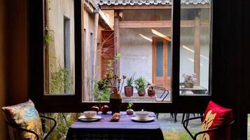 A dining area with a table set for two, a view of a courtyard through a window, and a chair with a cushion.
