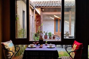 A dining area with a table set for two, a view of a courtyard through a window, and a chair with a cushion. - Yanshe Inn (Huangshan City)