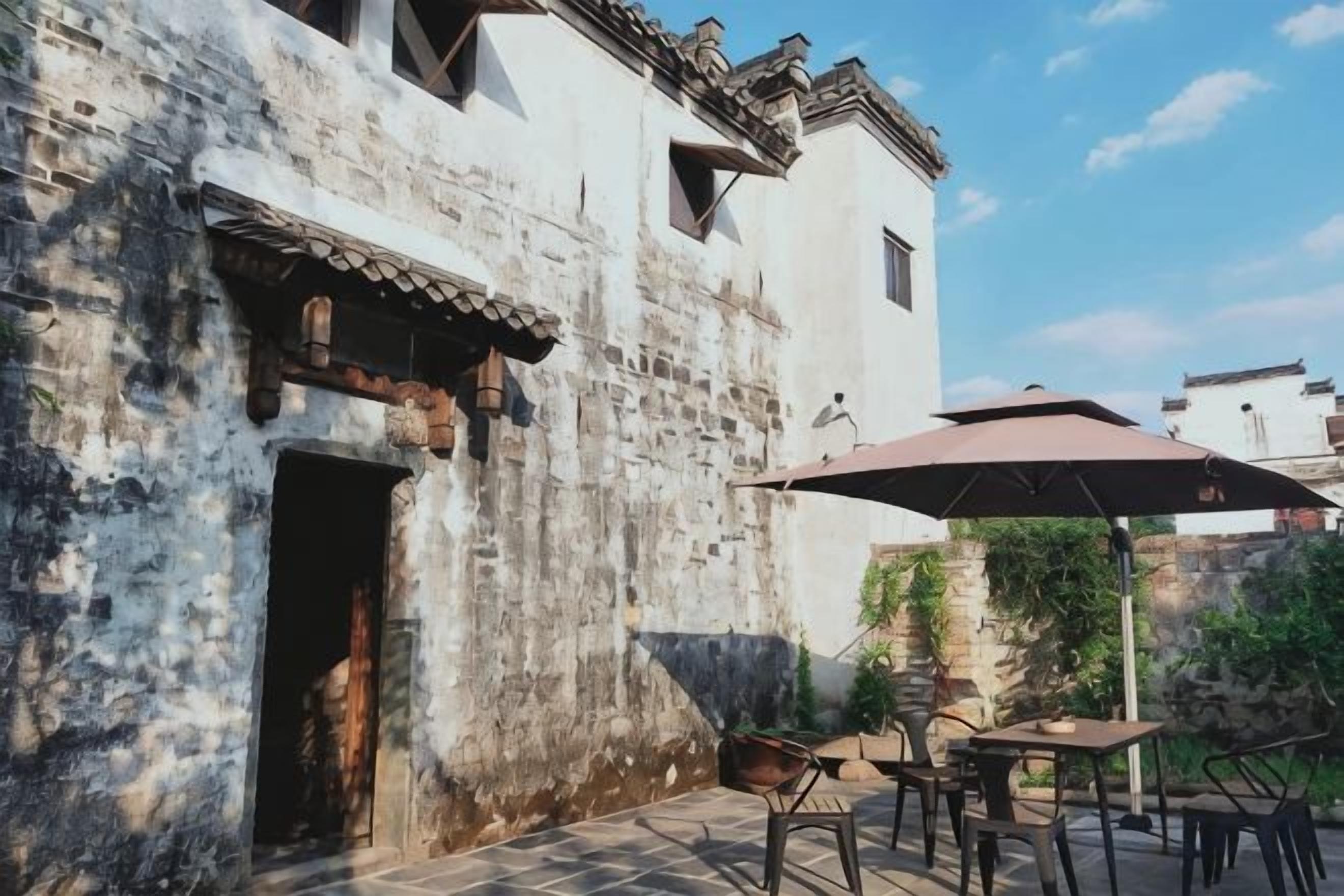 Un edificio rústico con un patio empedrado, mesas al aire libre y un gran paraguas que da sombra.