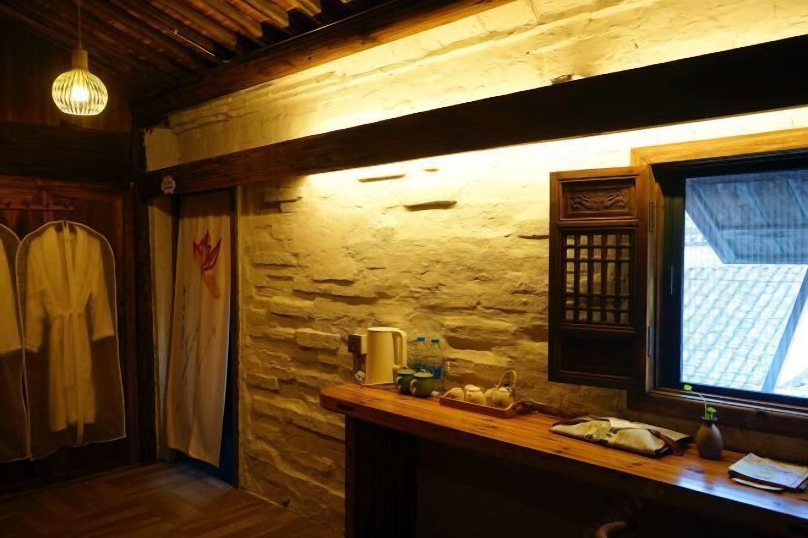 A rustic room with stone walls, wooden beams, a wooden table with a basket of apples, and a window with a view of a tiled roof.