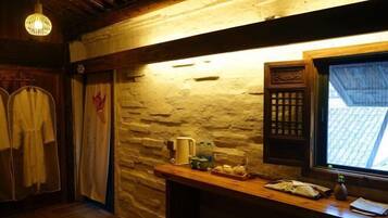 A rustic room with stone walls, wooden beams, a wooden table with a basket of apples, and a window with a view of a tiled roof.