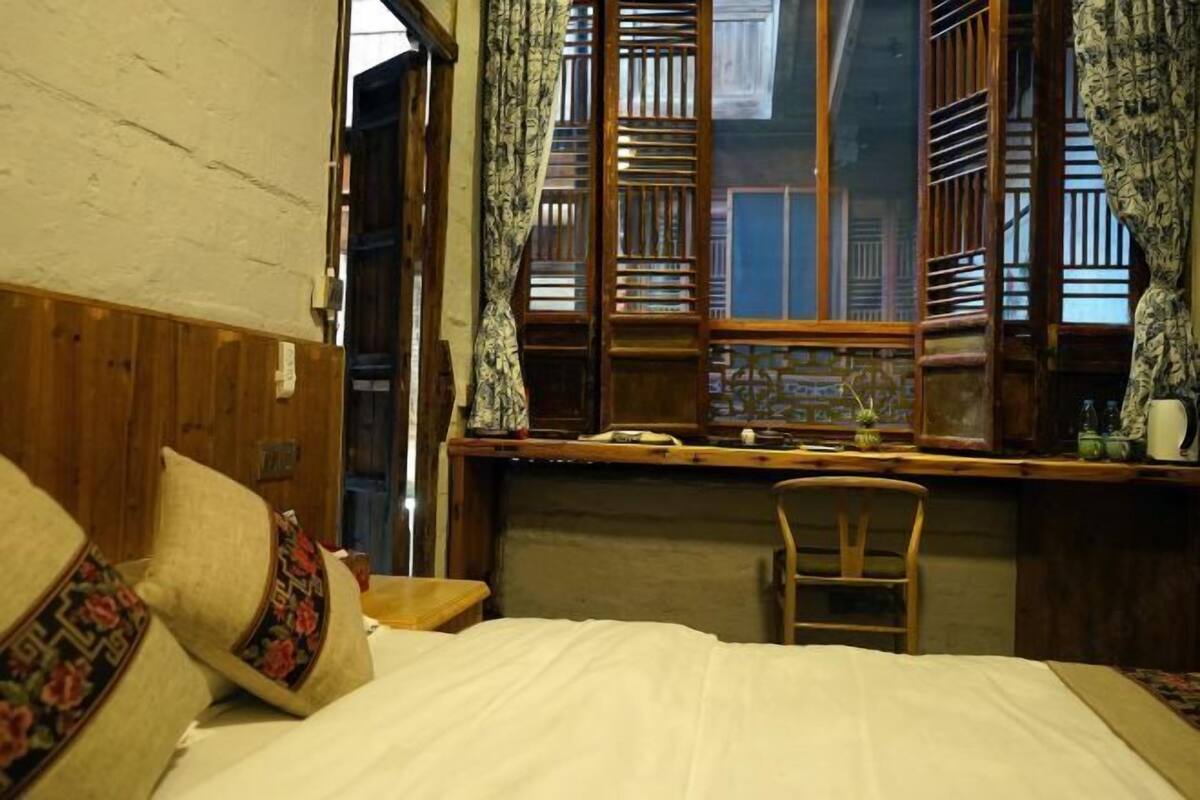 A room with a bed, wooden headboard, a window with wooden shutters, and a small table with a chair.