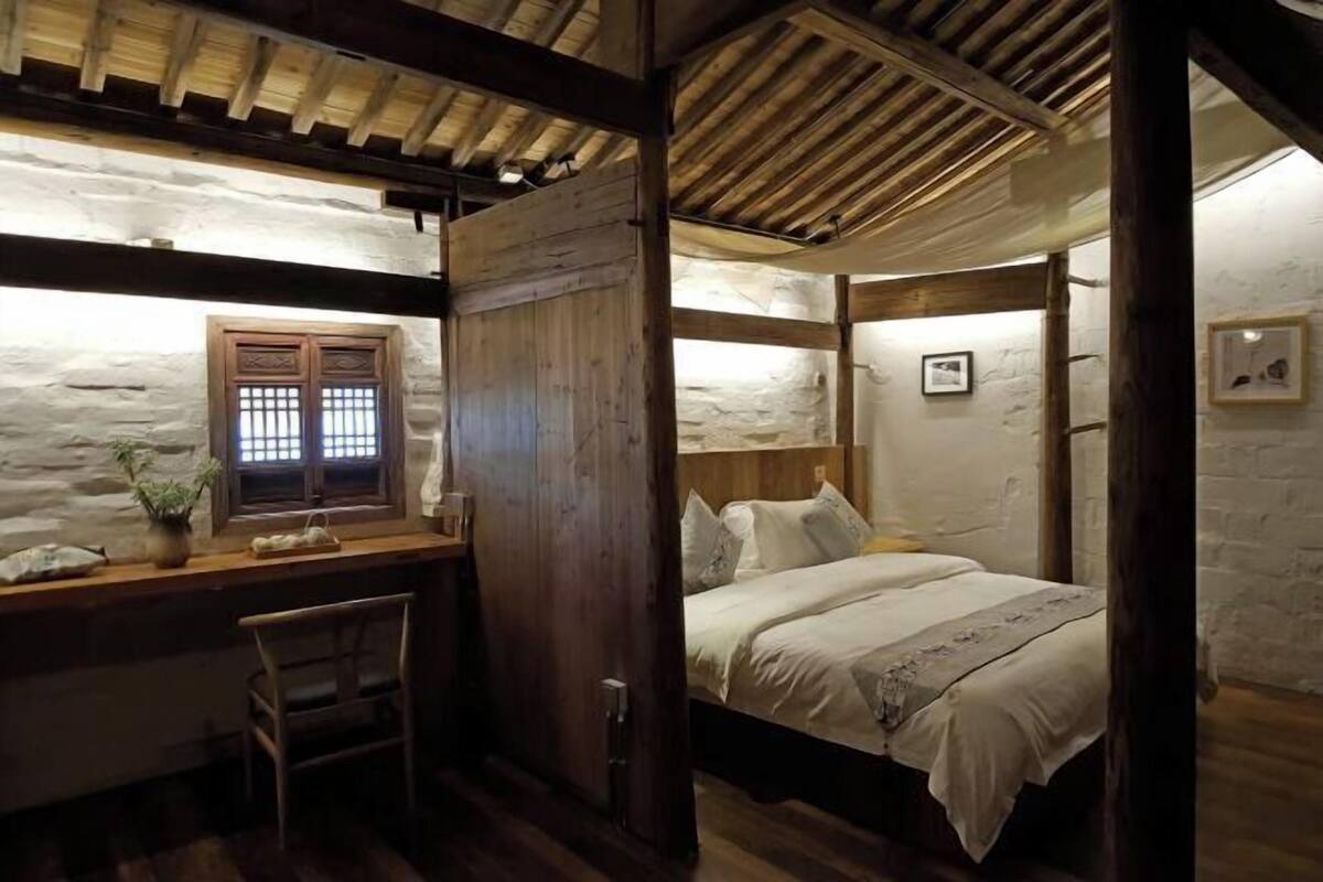 A rustic room with a wooden desk, a bed with white linens, and a stone wall.