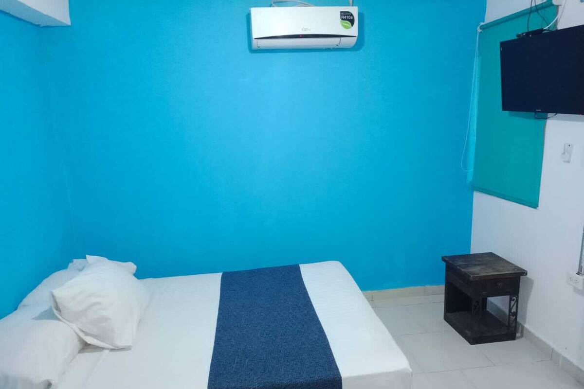Quarto com cama, mesinha, ar condicionado e televisão fixada na parede.