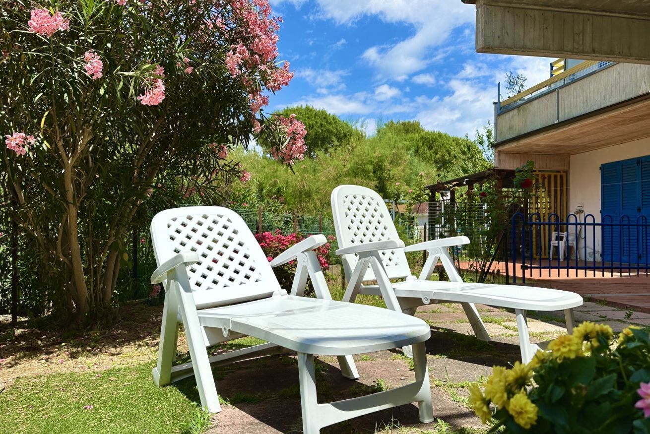 Two white lounge chairs in a garden with pink flowers and a building with a blue door in the background.