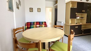 A dining area with a round wooden table and four chairs, a colorful sofa, and a kitchen area with cabinets and a radiator. - M341 - Marcelli, Three-room apartment with garden close to the sea (Marcelli)
