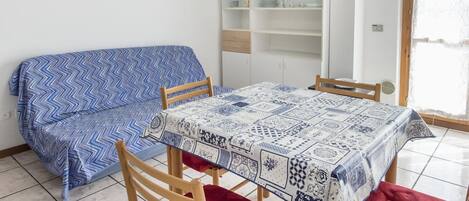 A room with a sofa, a table with a patterned tablecloth, a wooden chair, and a built-in shelving unit.