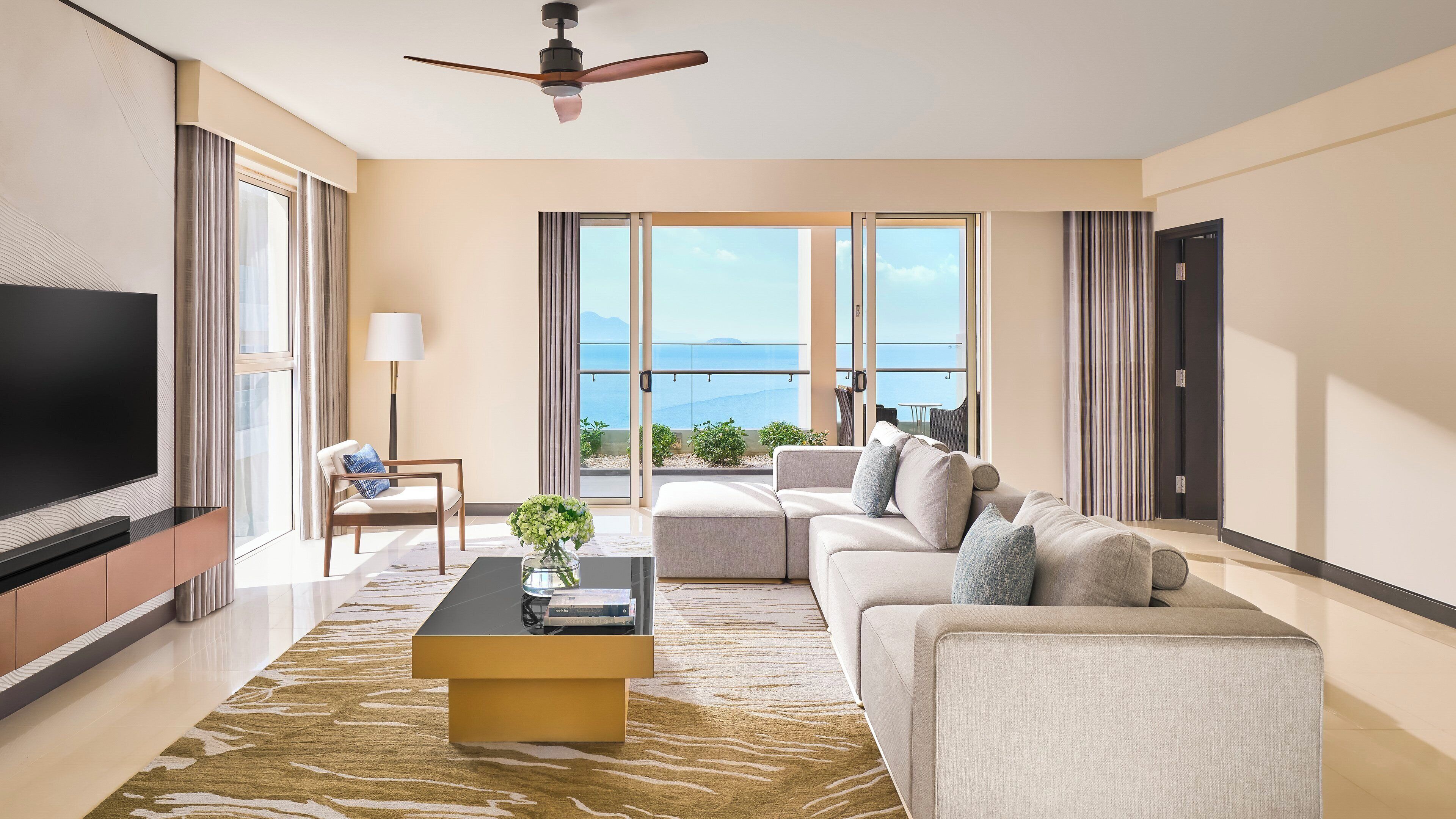 A modern living room with a flat-screen TV, a sofa, a coffee table, and a view of the sea through large windows.