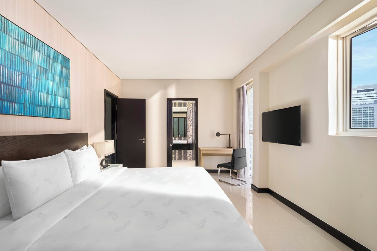 A modern hotel room with a large bed, a desk with a chair, a TV, and a city view.