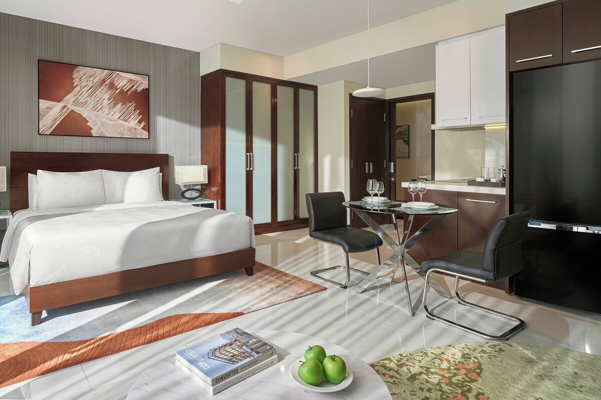 A hotel room with a large bed, a desk with a chair, a dining table with chairs, a kitchen area, and a bathroom.