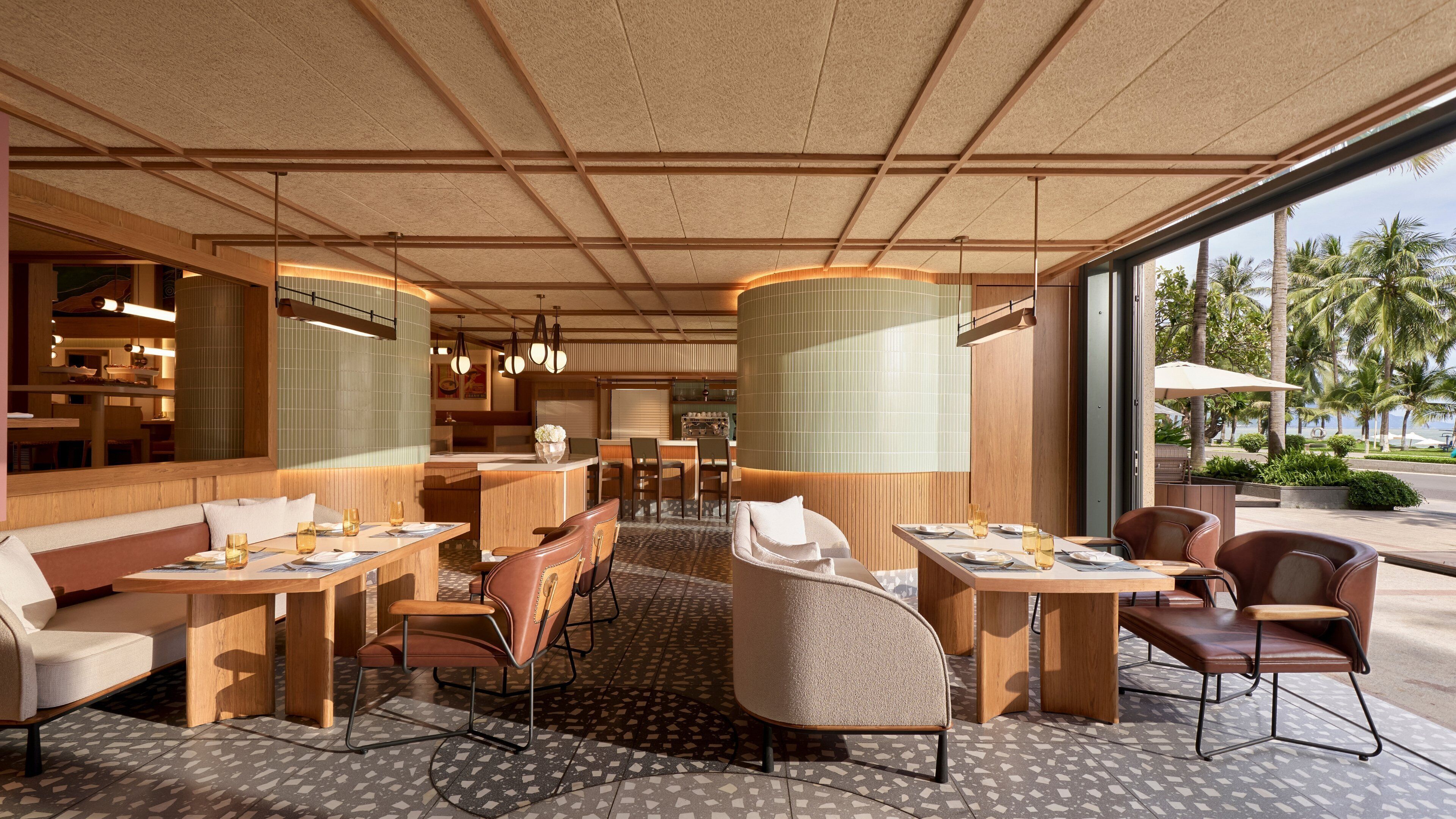 A modern restaurant with a mix of indoor and outdoor seating, featuring a central island and a view of palm trees.