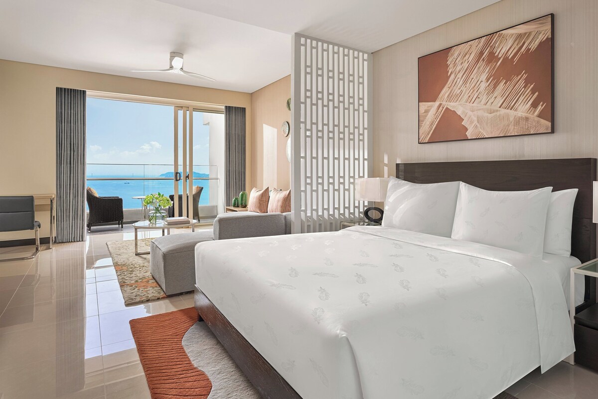 A hotel room with a large bed, a bedside table with a lamp, a view of the sea, and a balcony with a chair.