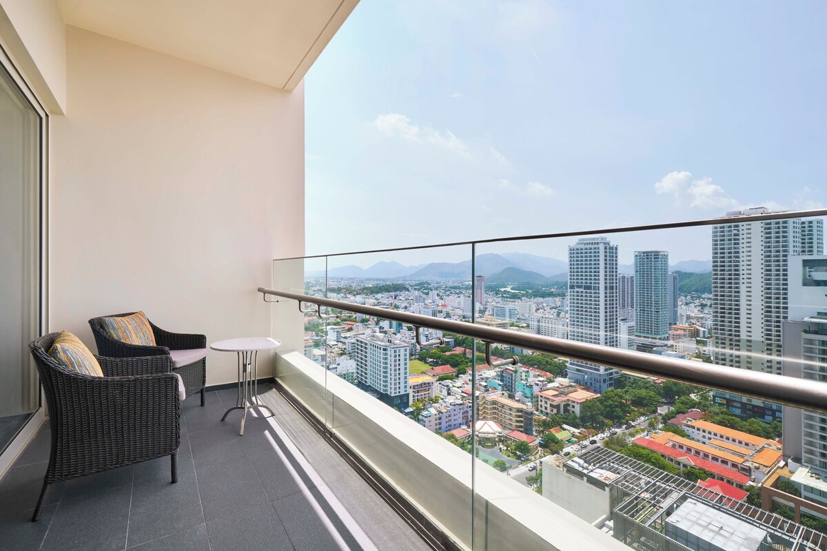 A balcony with wicker chairs and a small table, offering a cityscape view with tall buildings and mountains in the background.