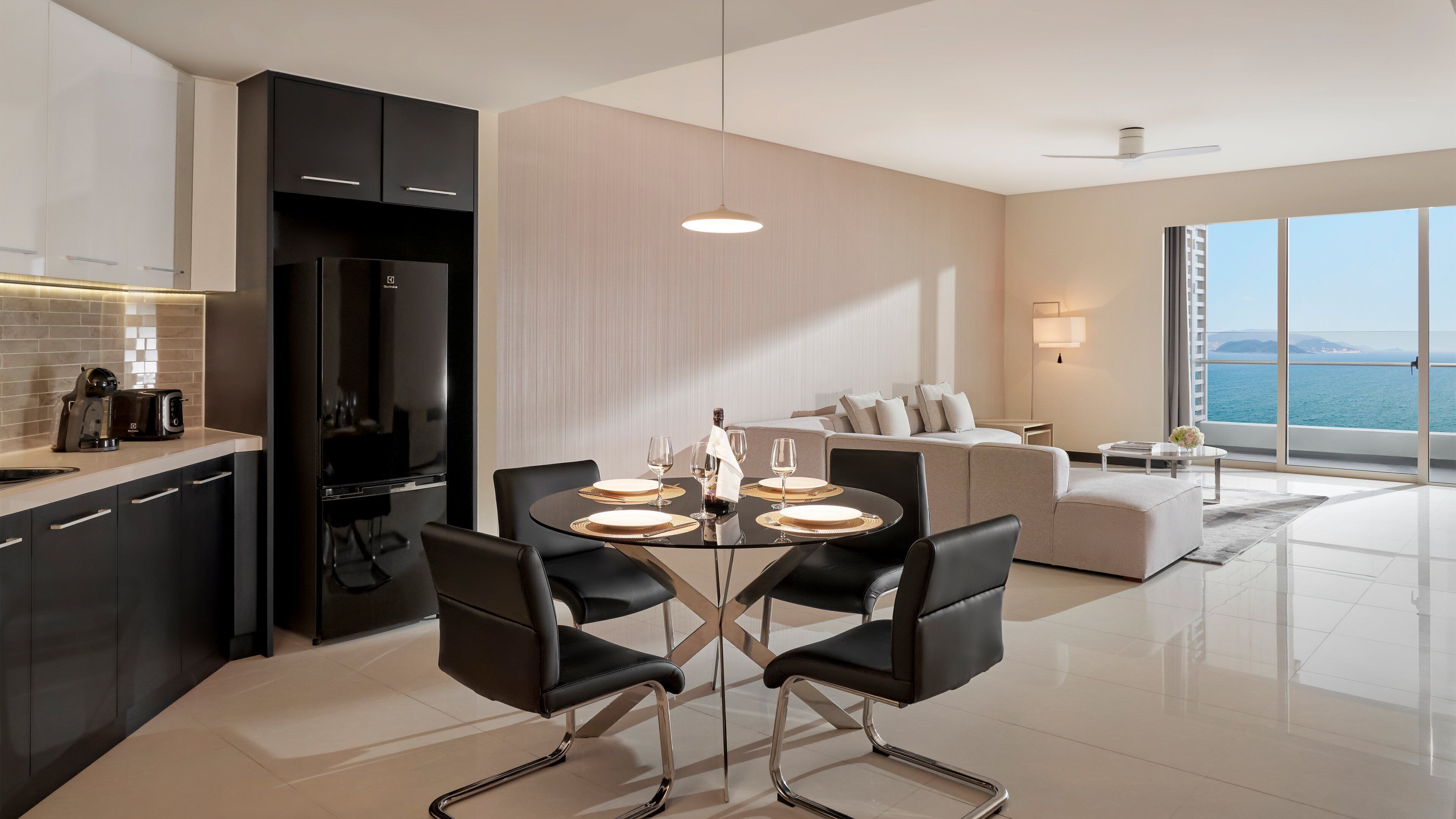 A modern hotel room with a dining area, kitchen, and a balcony with a view of the sea.