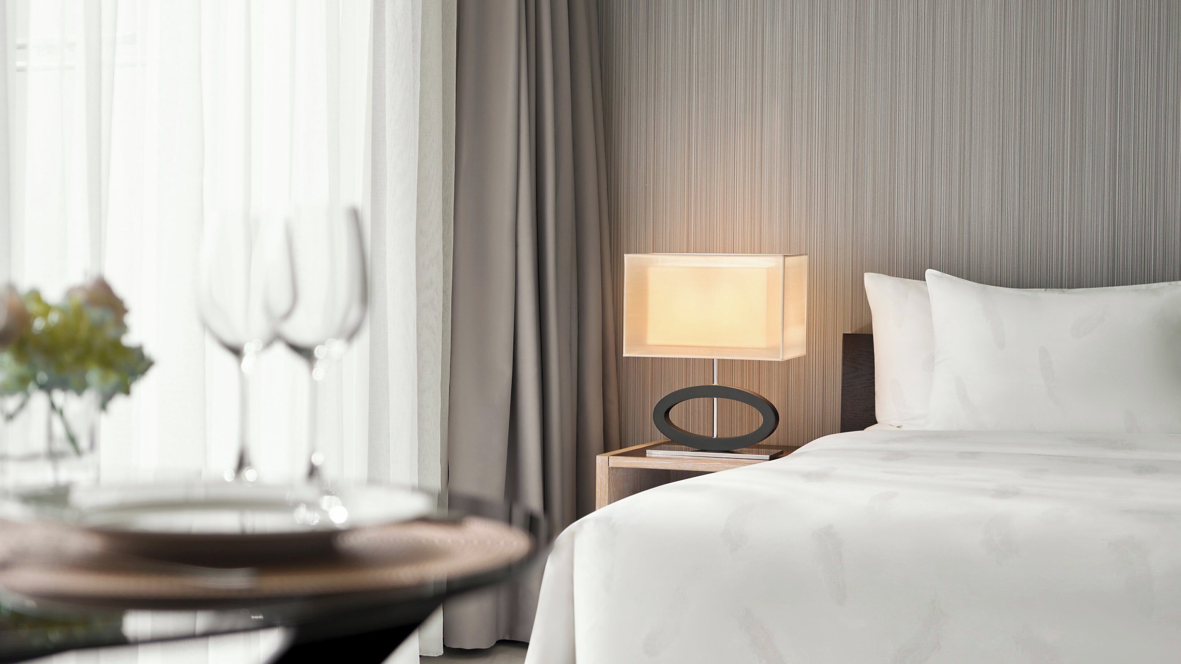 A hotel room with a bed, a bedside table with a lamp, and a dining area with a table and chairs.