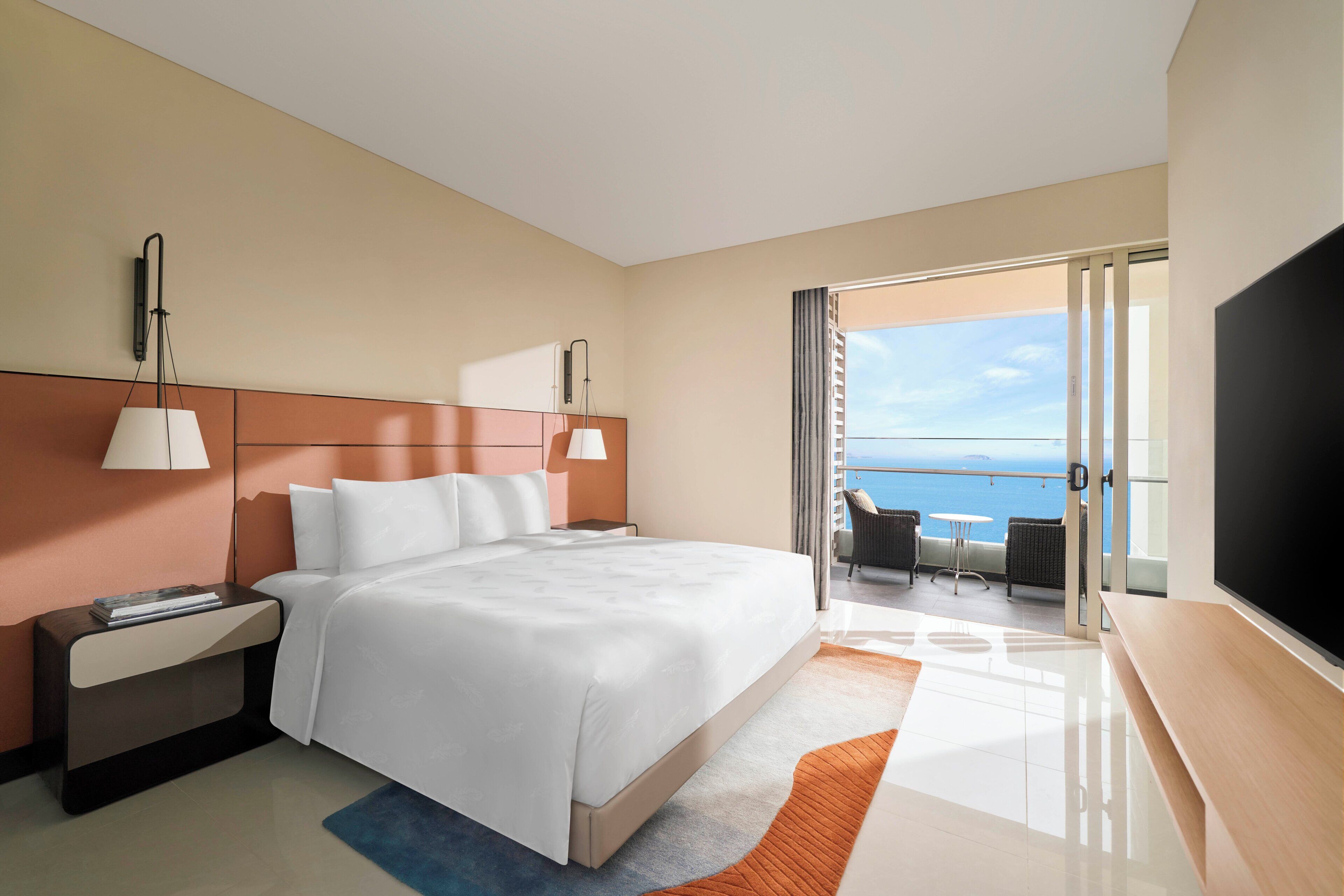A hotel room with a large bed, a TV, a desk, and a balcony with a view of the sea.