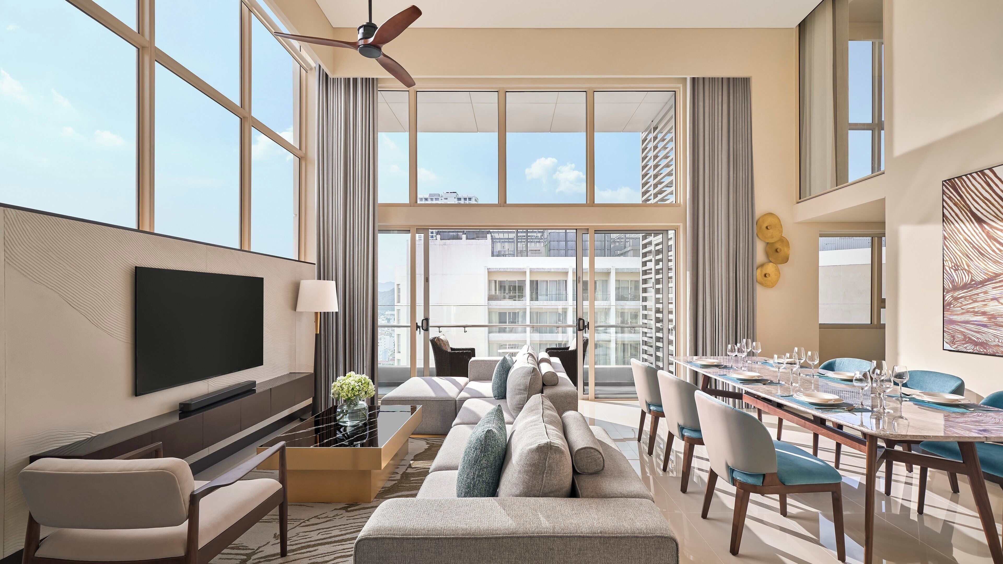 A modern hotel room with a large TV, comfortable seating, a dining area, and large windows offering city views.