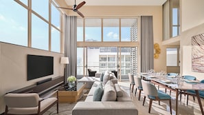 Una habitación de hotel moderna con un televisor grande, asientos cómodos, una zona de comedor y grandes ventanales con vistas a la ciudad.