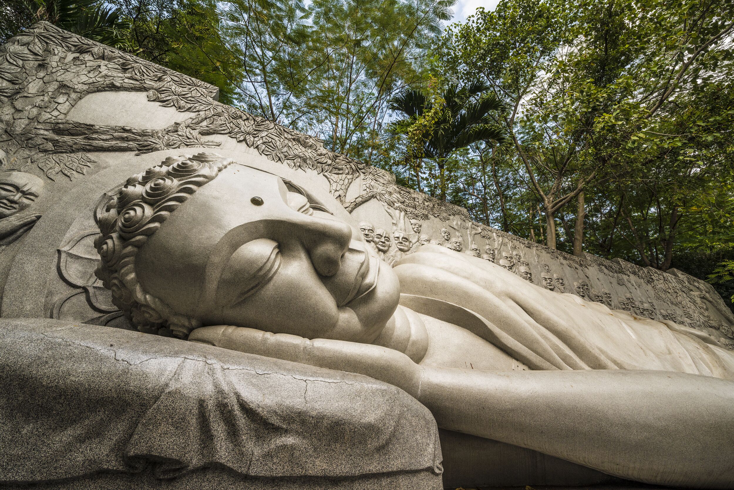 A large stone statue of a reclining figure with intricate carvings, set against a backdrop of trees.