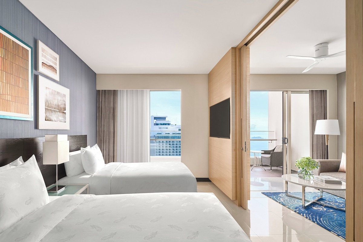 A hotel room with two beds, a large window with a city view, a balcony with seating, and a flat-screen TV.