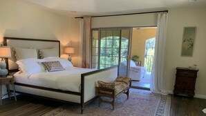 Quarto com uma cama grande, uma cadeira, mesa de cabeceira, luminária, janela com cortinas e vista para o exterior.