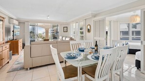A bright, open-plan living area with a glass dining table, white chairs, a beige sofa, and a kitchen area with wooden cabinets. - The Moroccan - Q Stay (Surfers Paradise)