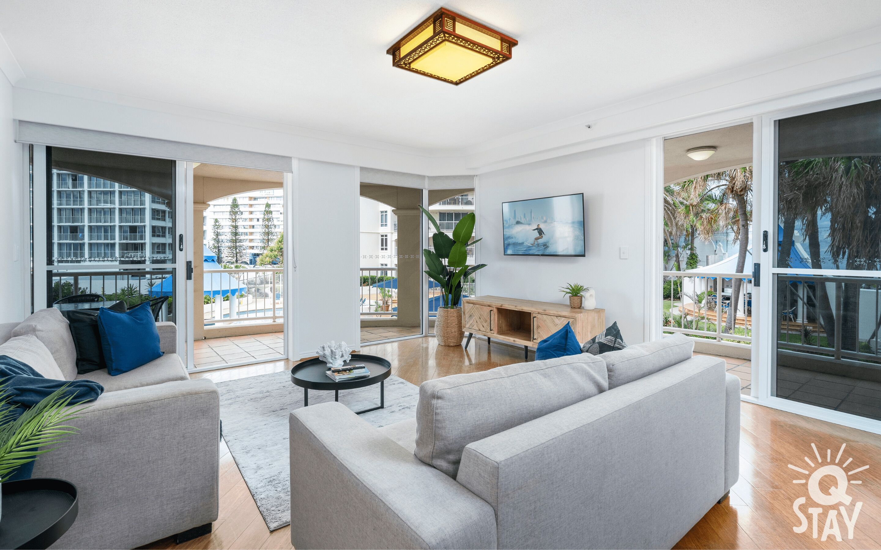 A spacious living room with a grey sofa set, a coffee table, and a large window offering a view of a balcony and outdoor area.