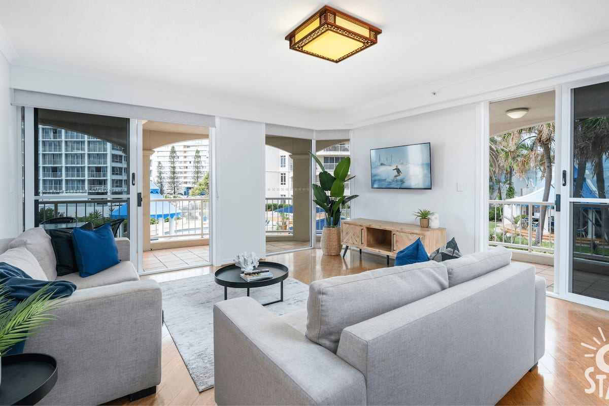 A spacious living room with a grey sofa set, a coffee table, and a large window offering a view of a balcony and outdoor area.