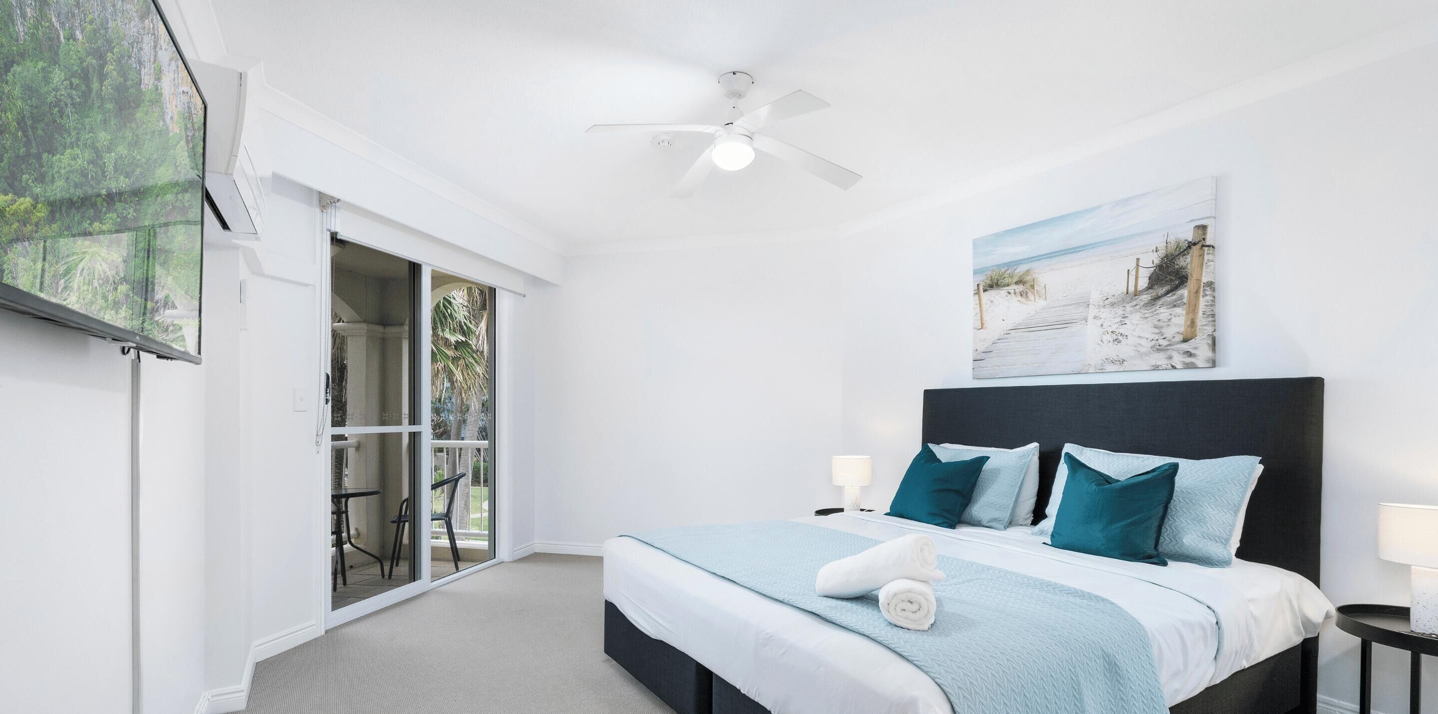 A bedroom with a bed, a ceiling fan, a painting, and a view through a glass door.