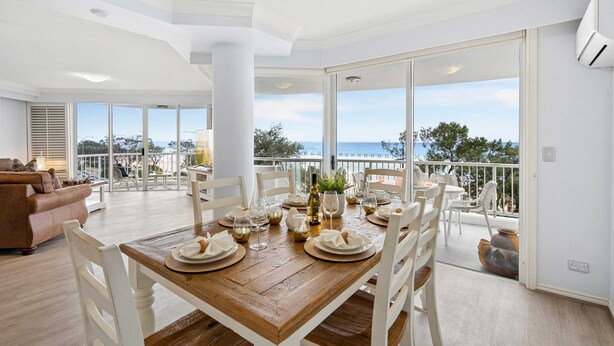 A spacious dining area with a wooden table set for a meal, white chairs, and a view of the sea through large windows. - The Moroccan - Q Stay (Surfers Paradise)