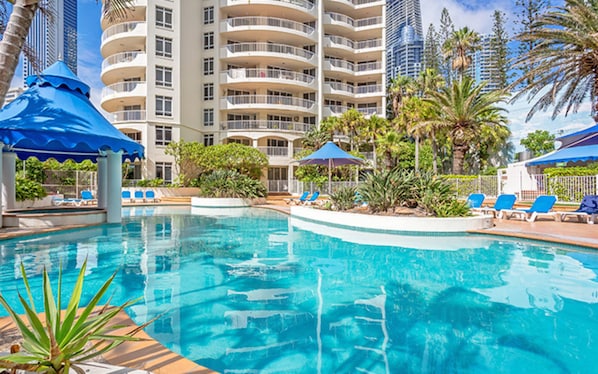 A swimming pool with blue umbrellas, lounge chairs, and palm trees.