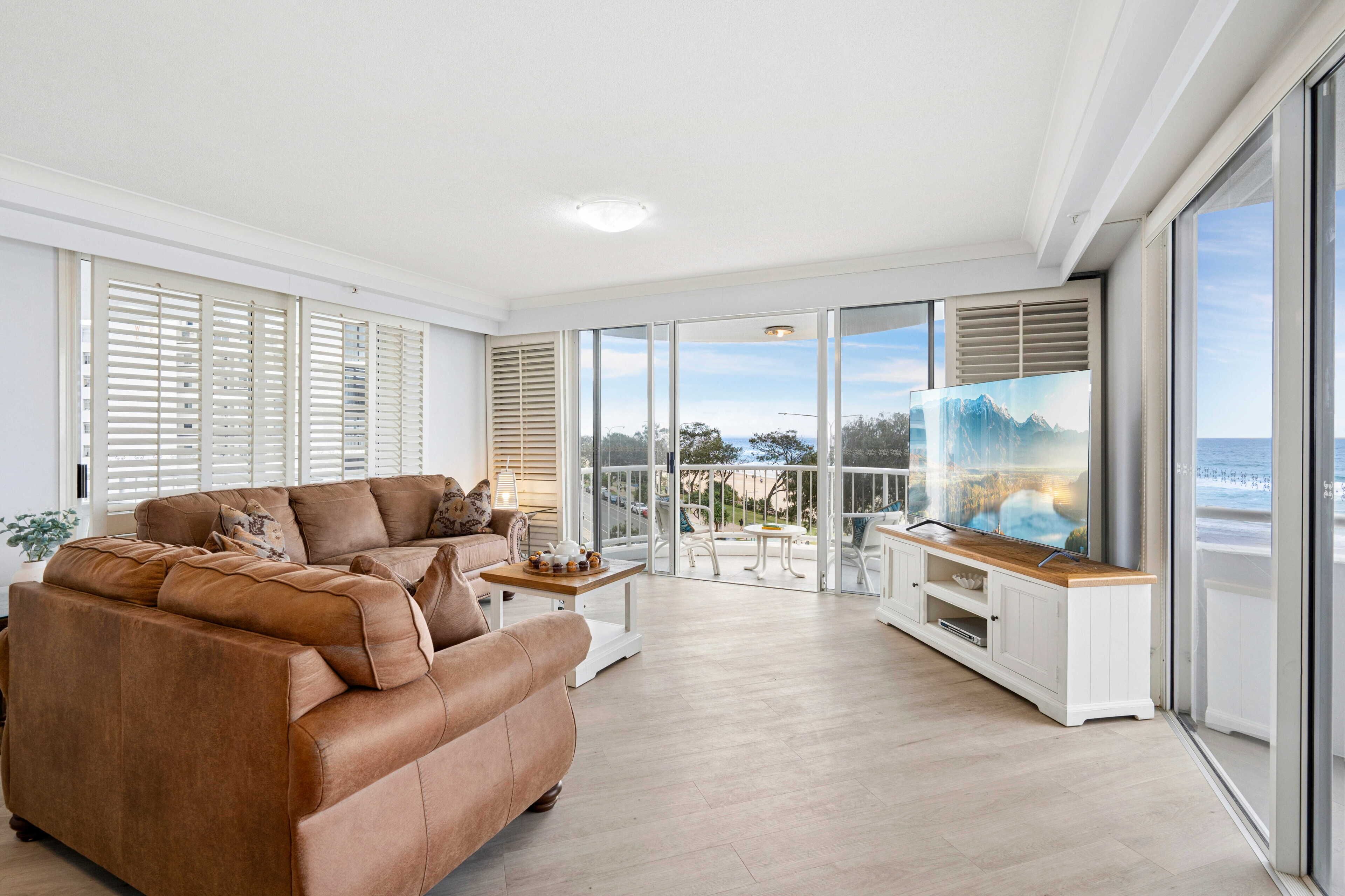 A spacious living room with a large sofa, a coffee table, and a TV. There are large windows with blinds, a balcony with outdoor furniture, and a view of the sea.