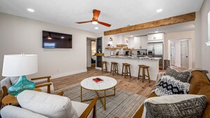 Una sala moderna con televisor, una mesa de centro y un sofá. La cocina tiene una isla central con taburetes y un ventilador de techo.