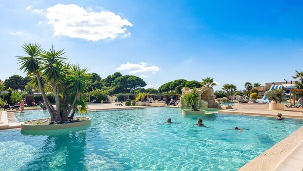 A swimming pool with clear blue water, surrounded by palm trees and rocks. - Mobil-home VIP | Clim & Plancha | Plage à 700 m (Vendres)
