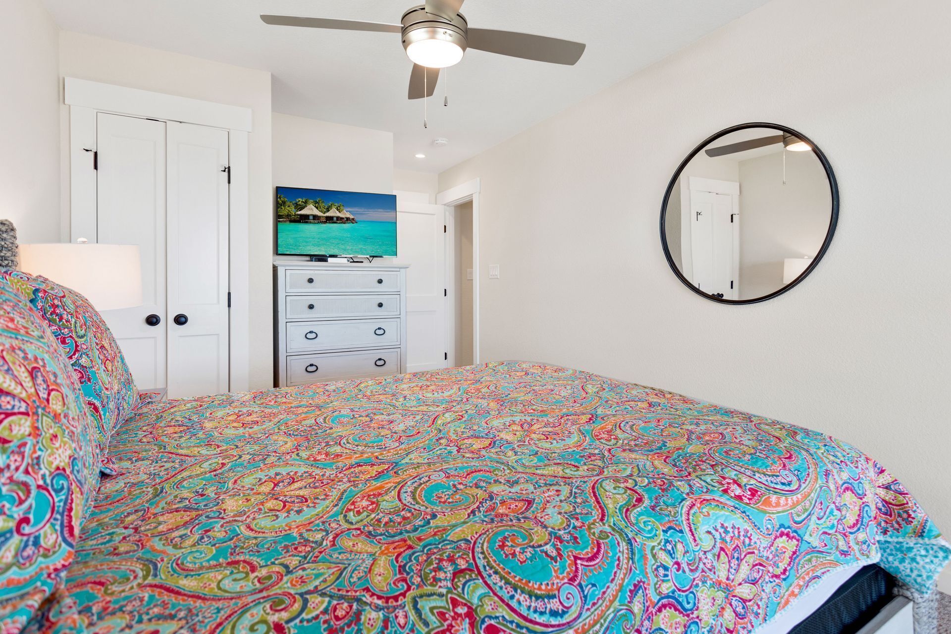 Une chambre à coucher avec une couvre-lit aux couleurs vives, un ventilateur de plafond, une commode, un tableau et un miroir rond.